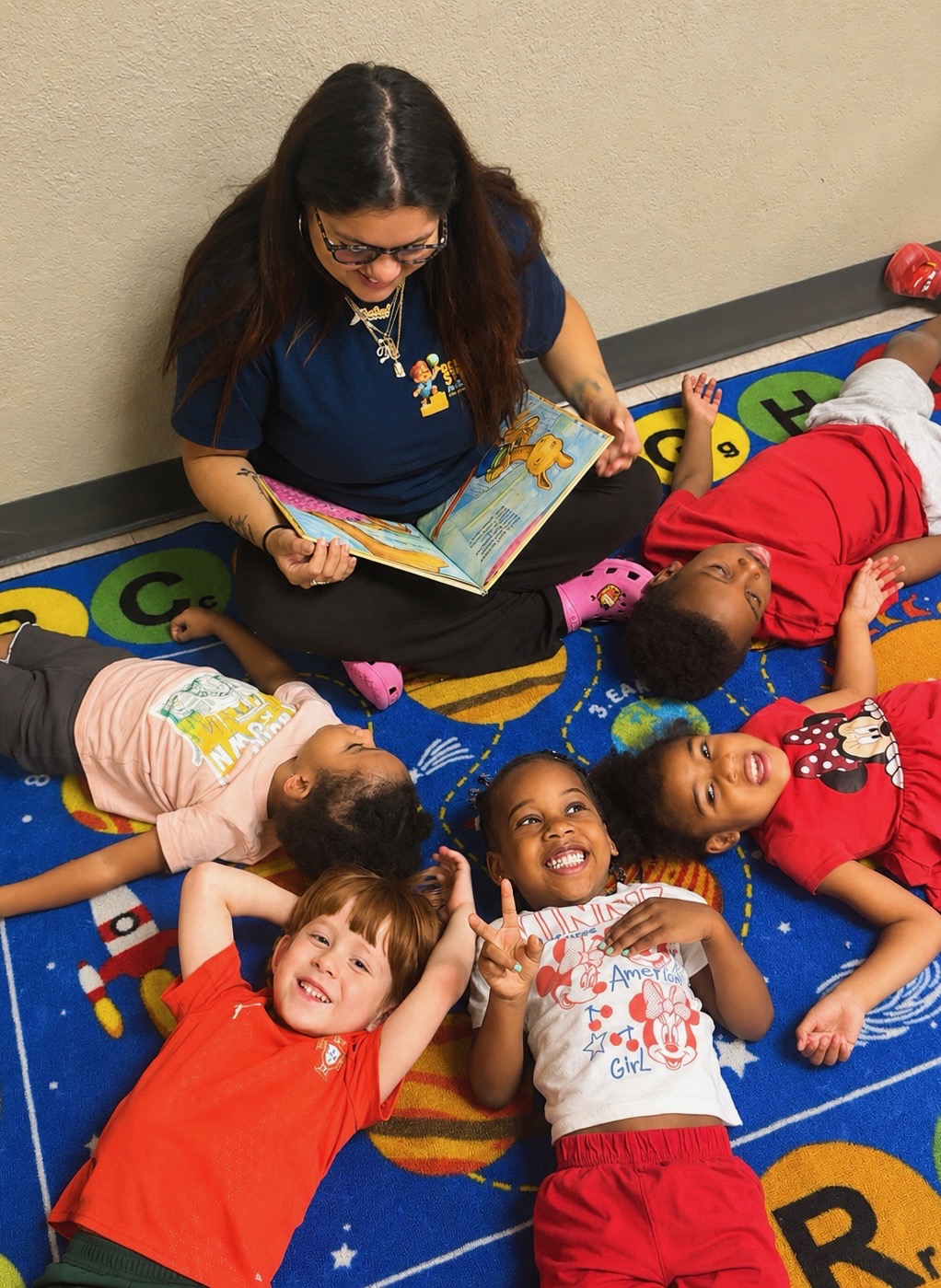 Teacher sitting on the classroom rug reading a book to a circle of smiling children